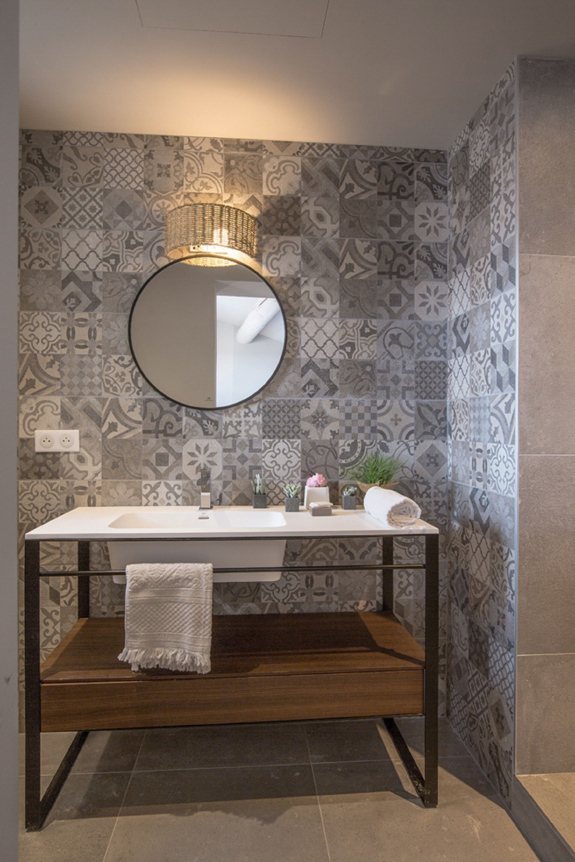 A bathroom with patterned tiles, a round mirror, a white sink, a towel on the counter, and a small plant.