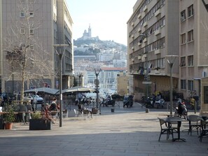 An outdoor café area with tables and chairs, surrounded by buildings and a view of a hill with a tower. - T2, Panier district, Old port, MuCEM (Marseille)