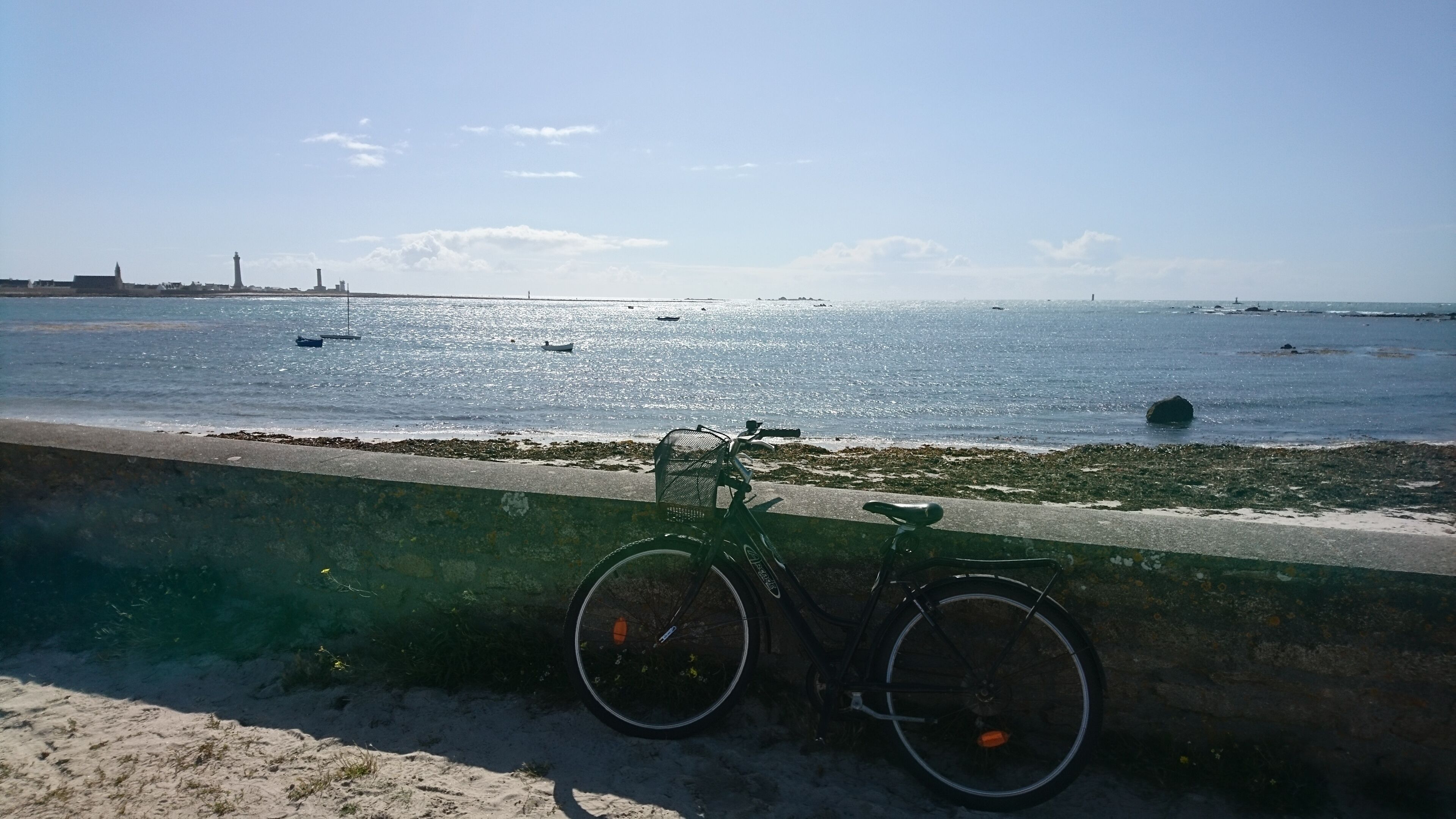 A bicycle parked by a concrete barrier overlooking a beach with boats and a distant lighthouse.