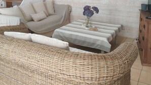 A cozy living room with a wicker sofa, a striped table, and a painting of kitchen utensils on the wall. - Pretty, well-equipped house, close to the sea and fishing port, ideal for children. (Penmarch)