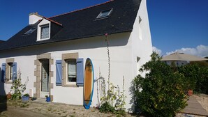 A white building with a black roof, blue shutters, and a surfboard leaning against the wall. - Pretty, well-equipped house, close to the sea and fishing port, ideal for children. (Penmarch)