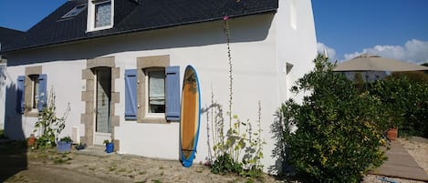 A white building with a black roof, blue shutters, and a surfboard leaning against the wall.