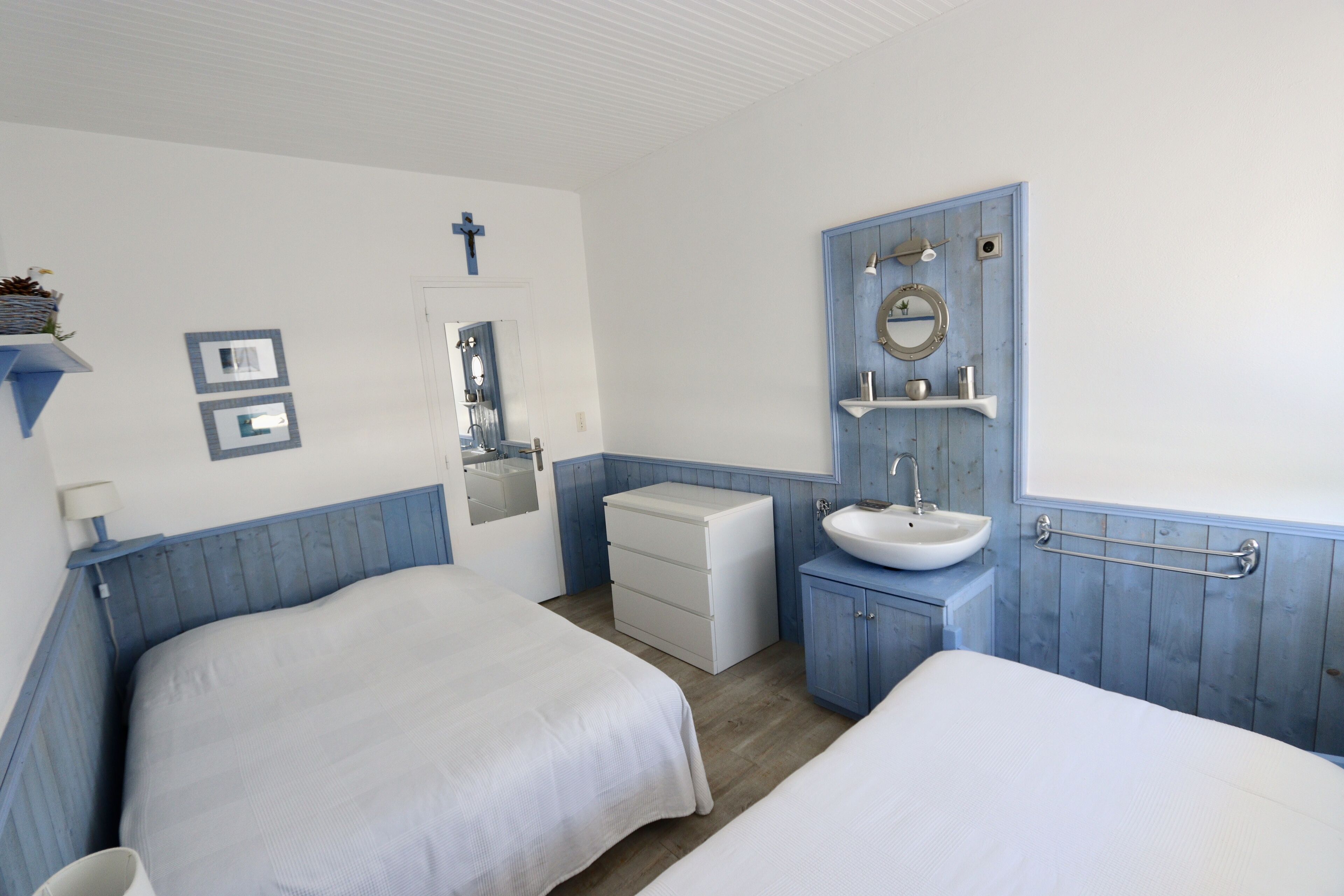 Habitación de hotel con dos camas, un lavamanos blanco, un espejo y una puerta con una cruz.