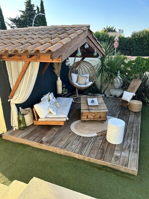 A cozy outdoor seating area with a wooden deck, a hanging chair, a cushioned bench, and a small table. - Air-conditioned villa with a private pool and hot tub, in the Camargue (Garons)