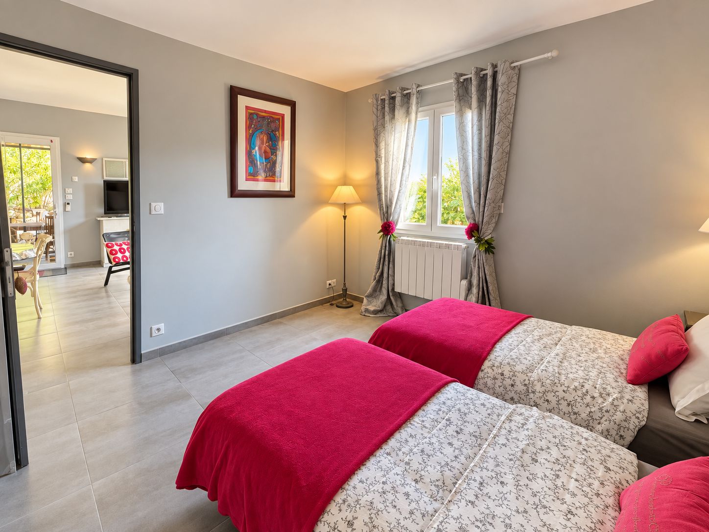 Quarto com duas camas, um quadro na parede, uma luminária e vista para o exterior.