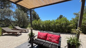 Ein überdachter Außenbereich mit einer Couch und Liegestühlen, umgeben von viel Grün und blauem Himmel.