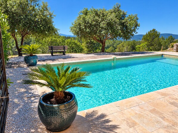 A swimming pool with a pebble border, surrounded by trees and a bench.