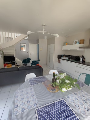 A modern kitchen with white cabinets, a dining area with patterned tablecloths, and a staircase leading to another level. - Charming house with seaside pool, WIFI, fully equiped kitchen, enclosed garden (Clohars-Carnoët)