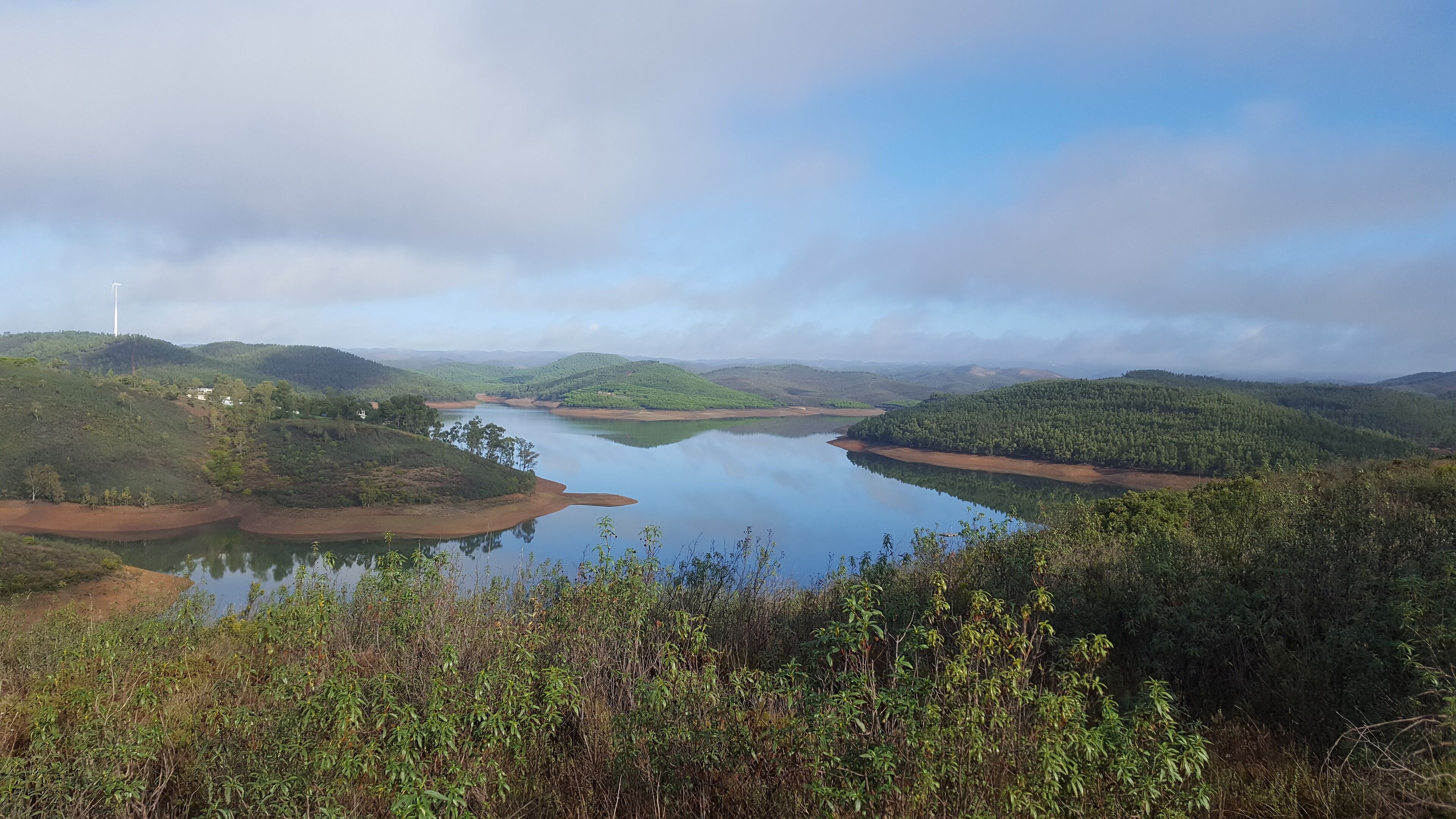 A panoramic view of a lake surrounded by hills with a wind turbine in the distance.