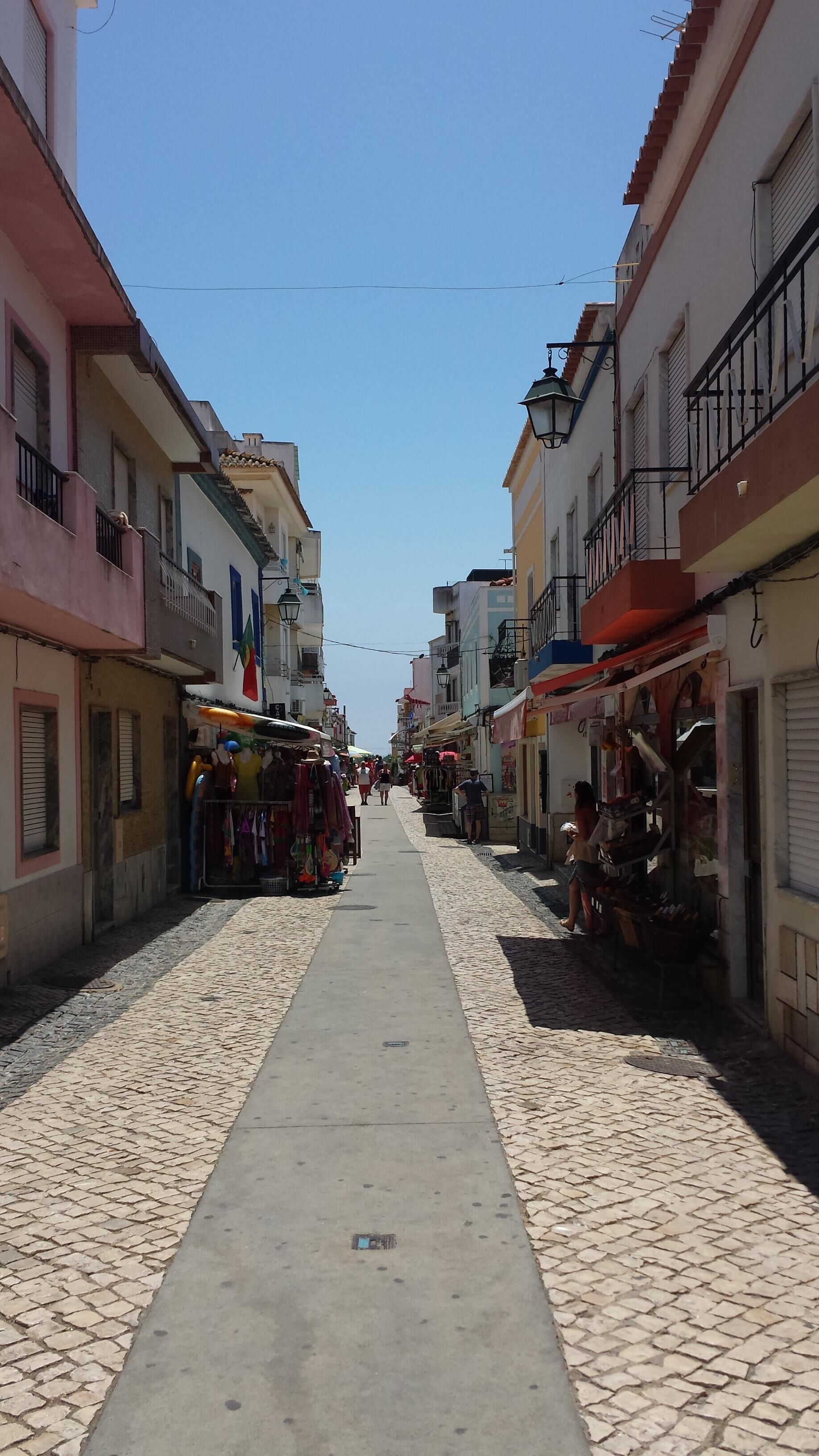 A narrow cobblestone street lined with buildings, some with balconies and shops on the ground floor.