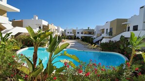 Une piscine entourée de bâtiments modernes dotés de balcons, le tout agrémenté de plantes et de fleurs tropicales.