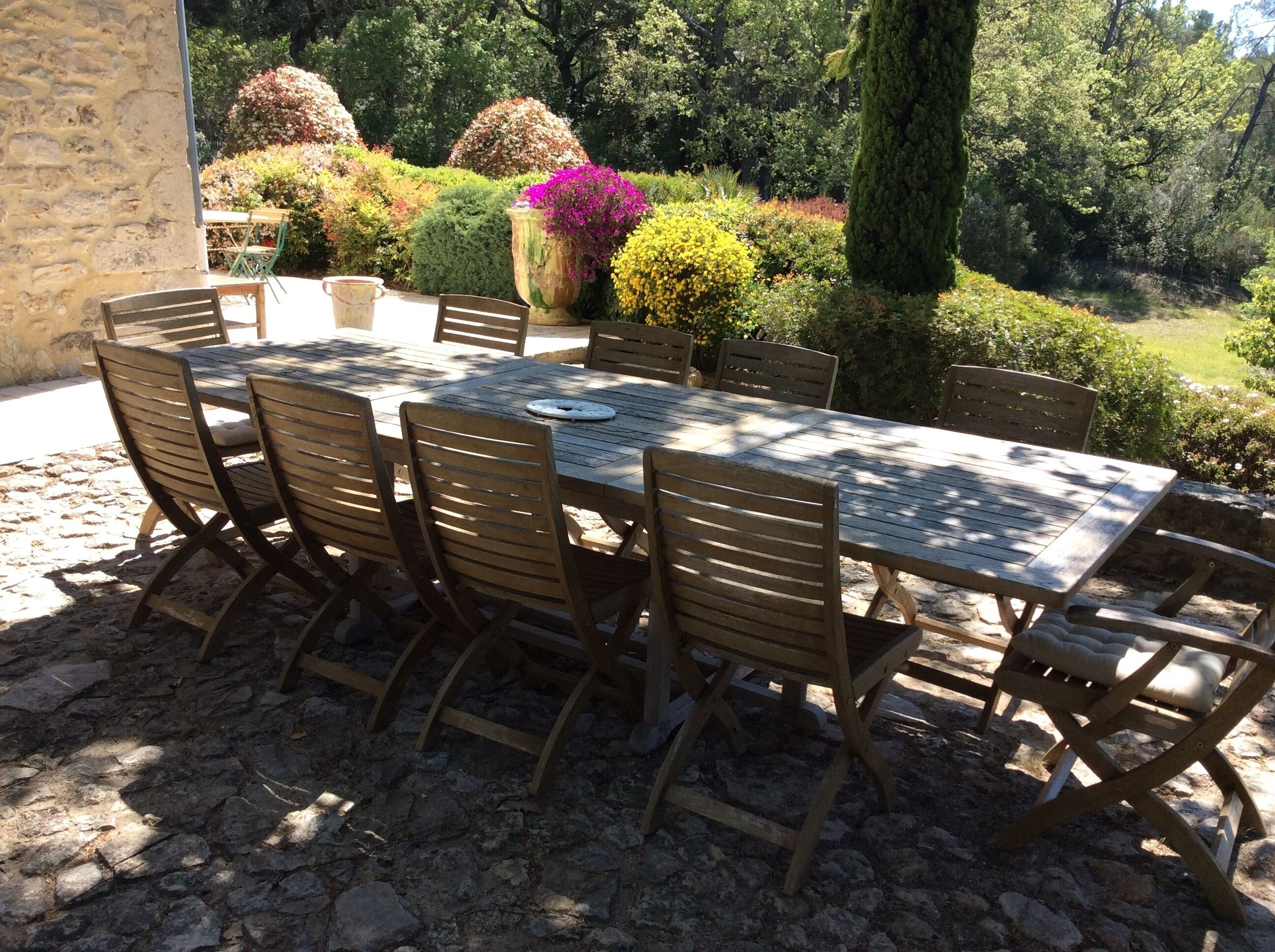 Un área de comedor al aire libre con un muro de piedra, una mesa con sillas y diversas plantas y árboles al fondo.