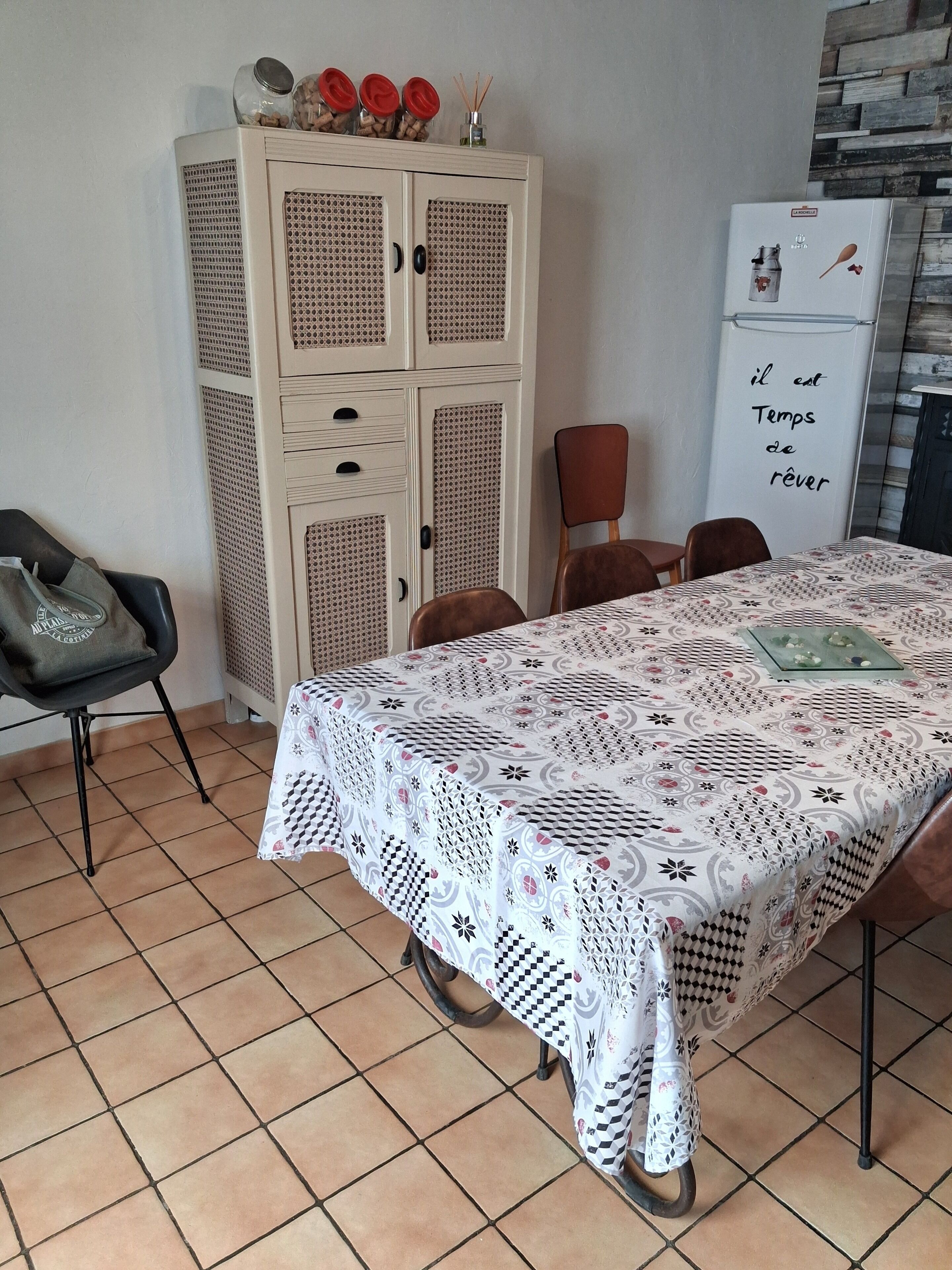 A dining area with a table covered in a patterned tablecloth, a cabinet with storage, a chair, and a refrigerator with a message on it.