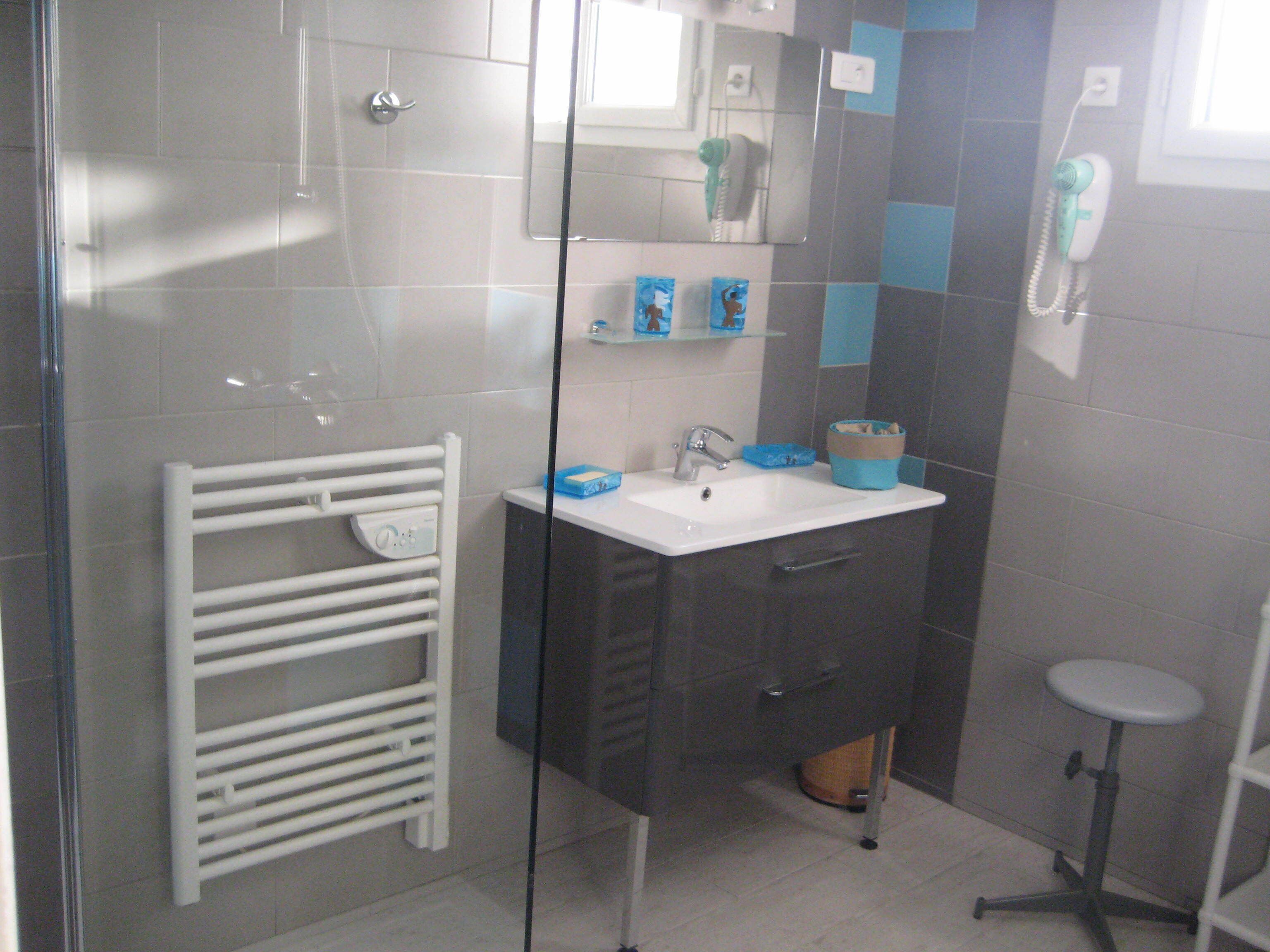 Un baño con cabina de ducha de vidrio, un toallero blanco, un lavamanos con espejo y un taburete pequeño.