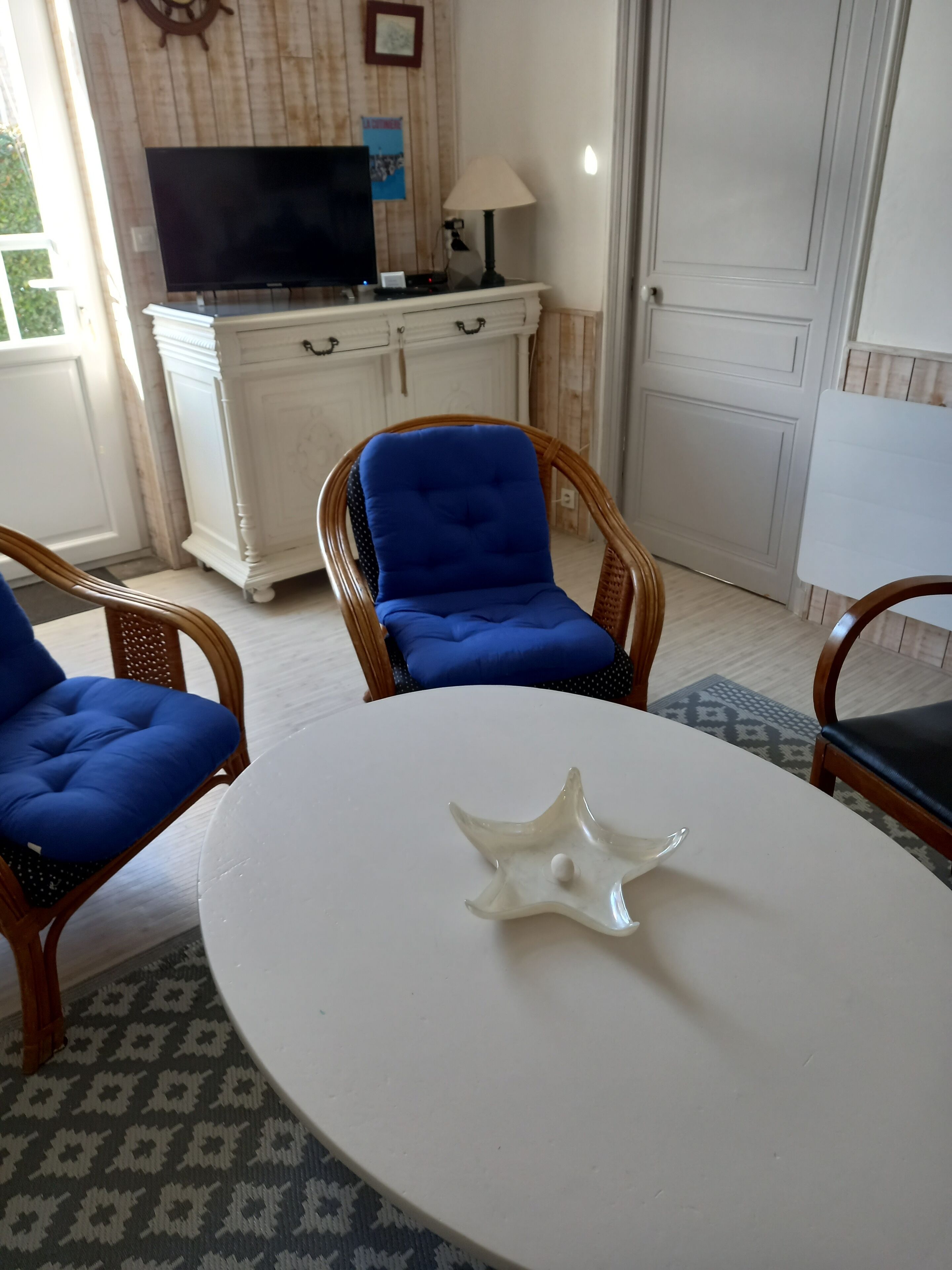 A room with a white round table, blue upholstered chairs, a white cabinet, and a television on a wooden wall.