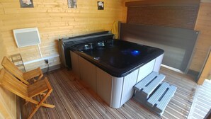 A small, cozy room with a wooden floor, a wooden bench, a hot tub, and a window with blinds.