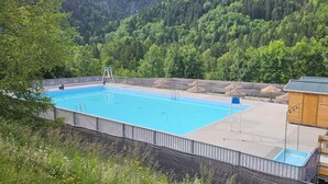 A swimming pool with a wooden deck, surrounded by a metal fence and a forest backdrop.