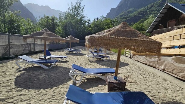A beach with sun loungers and thatched umbrellas, surrounded by wooden structures and mountains in the background.