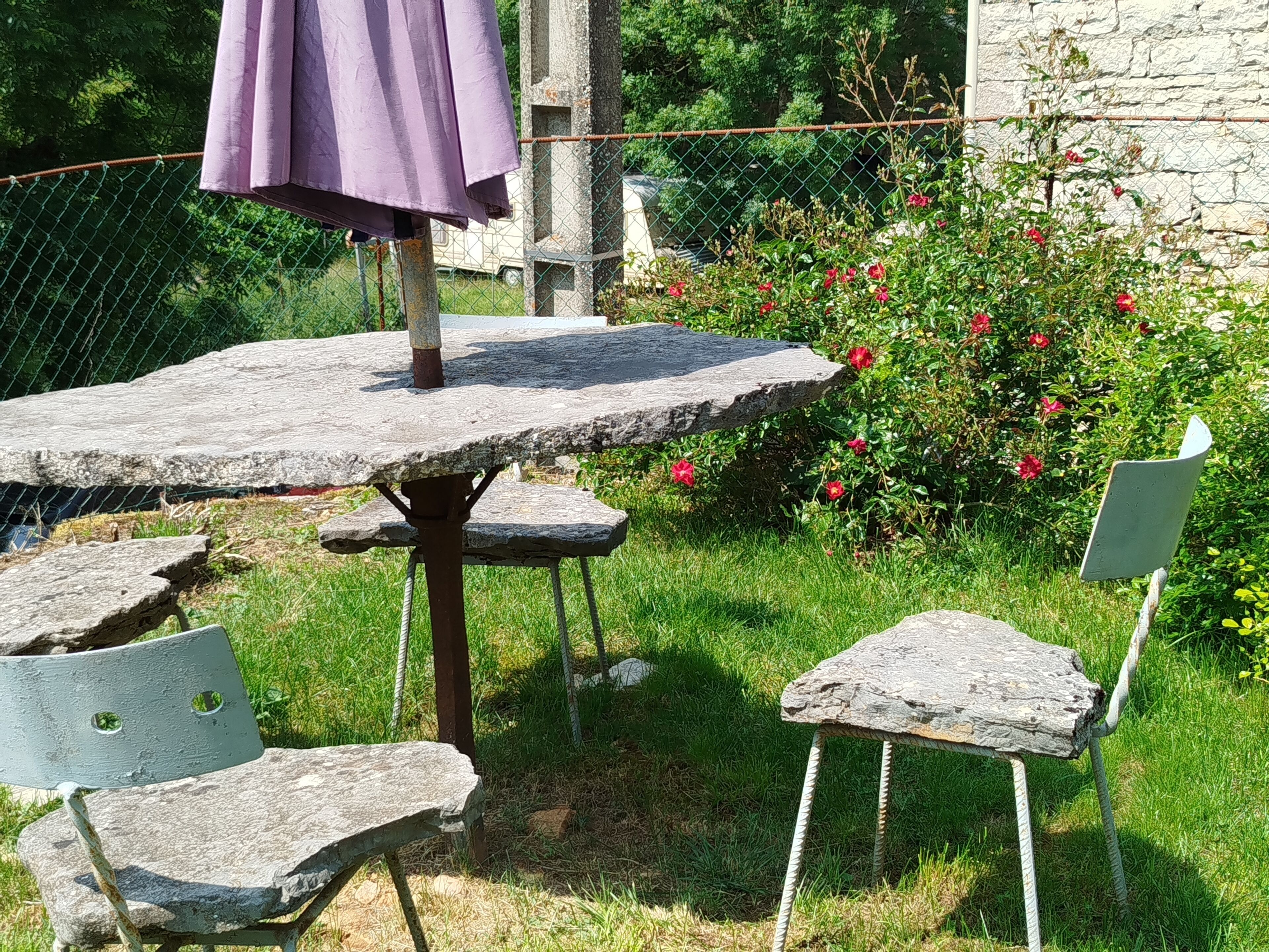 A stone table with metal chairs and a purple umbrella in a garden setting.
