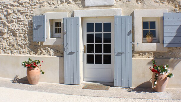 A stone building with a white door, two windows with shutters, and potted plants.