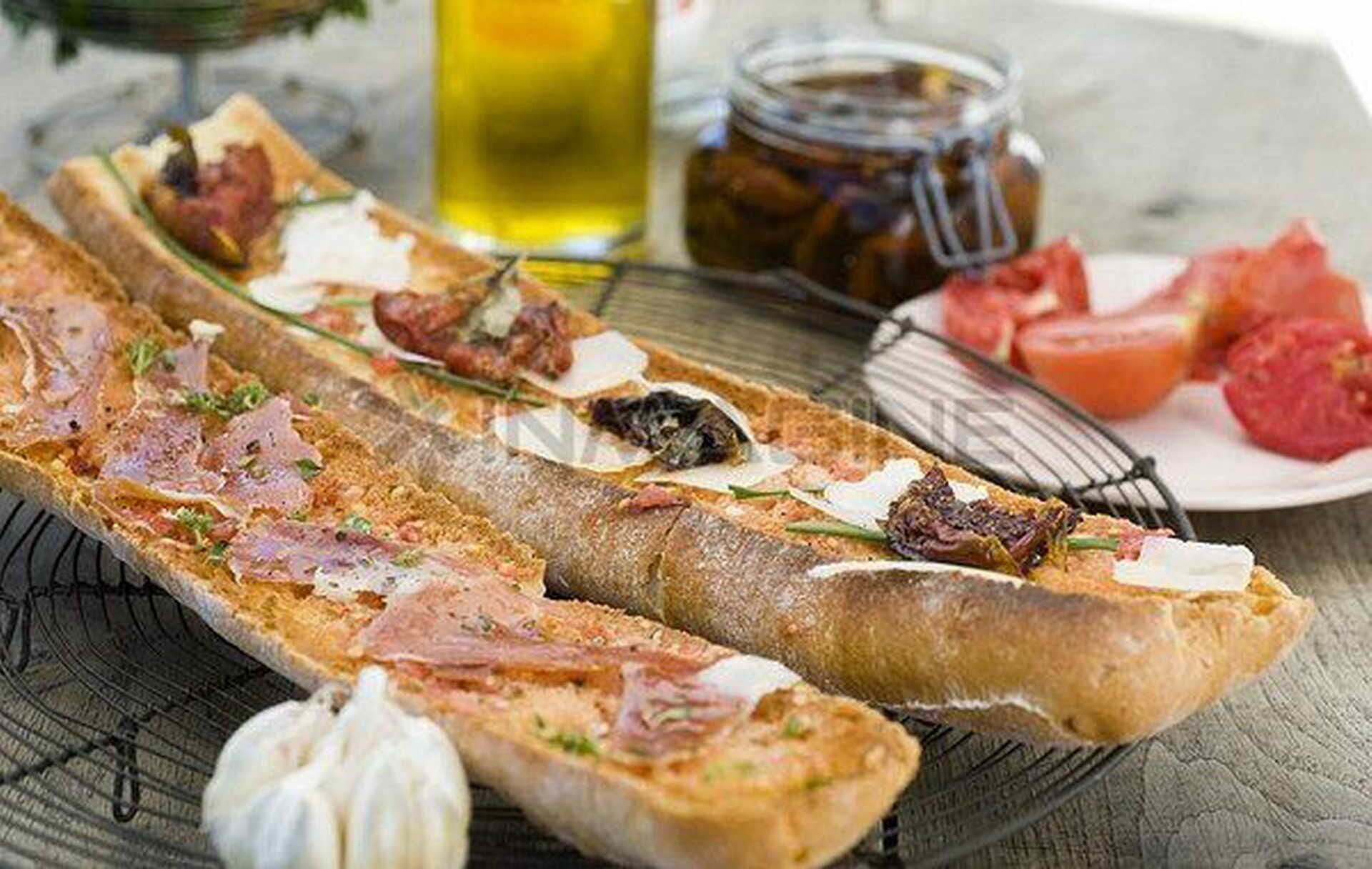 Une longue baguette, garnie de divers ingrédients, un petit bol d’ail et un bocal de cornichons sur une table.