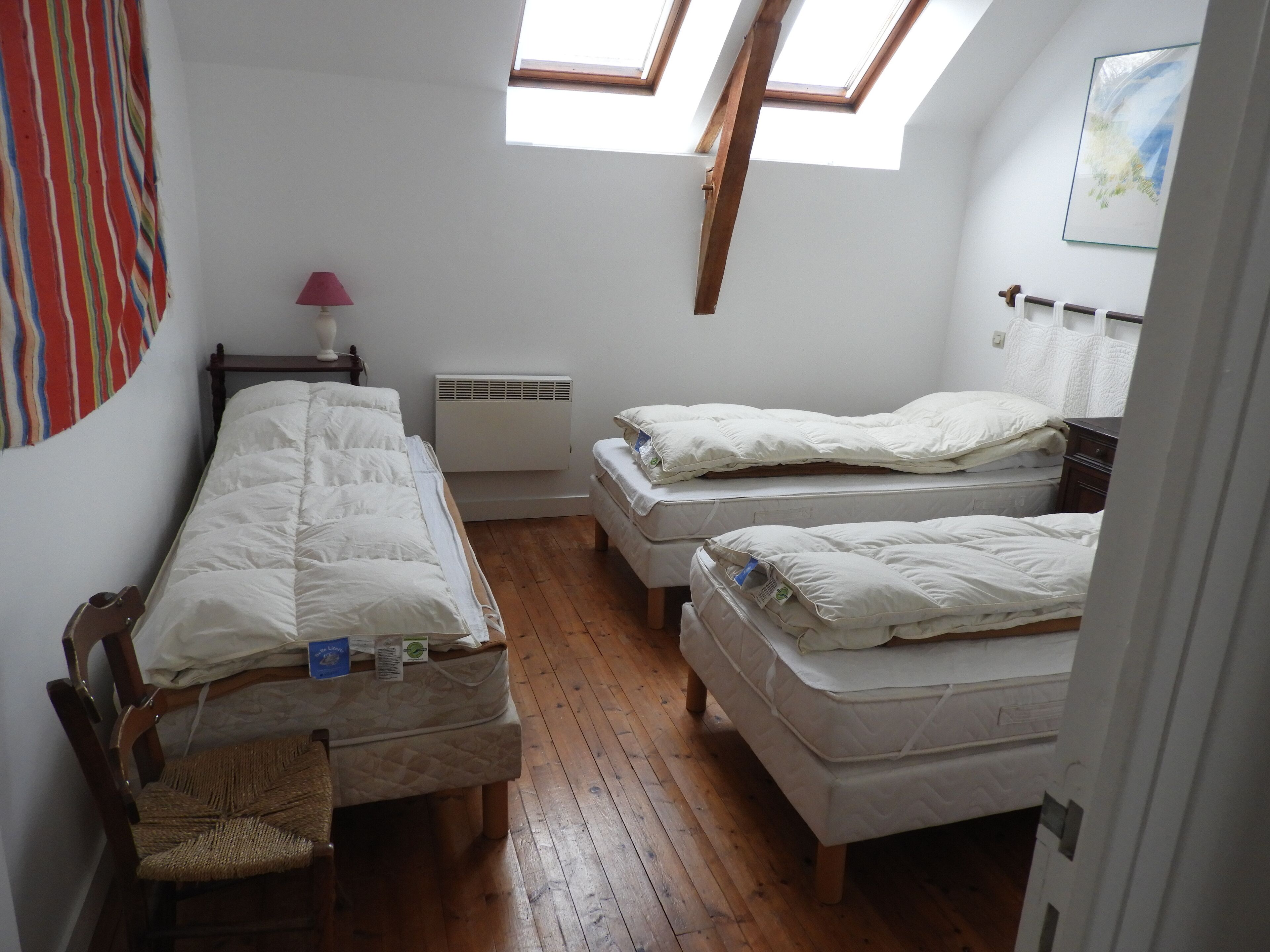 A dormitory room with two single beds, a wooden chair, a desk, a lamp, and a radiator.