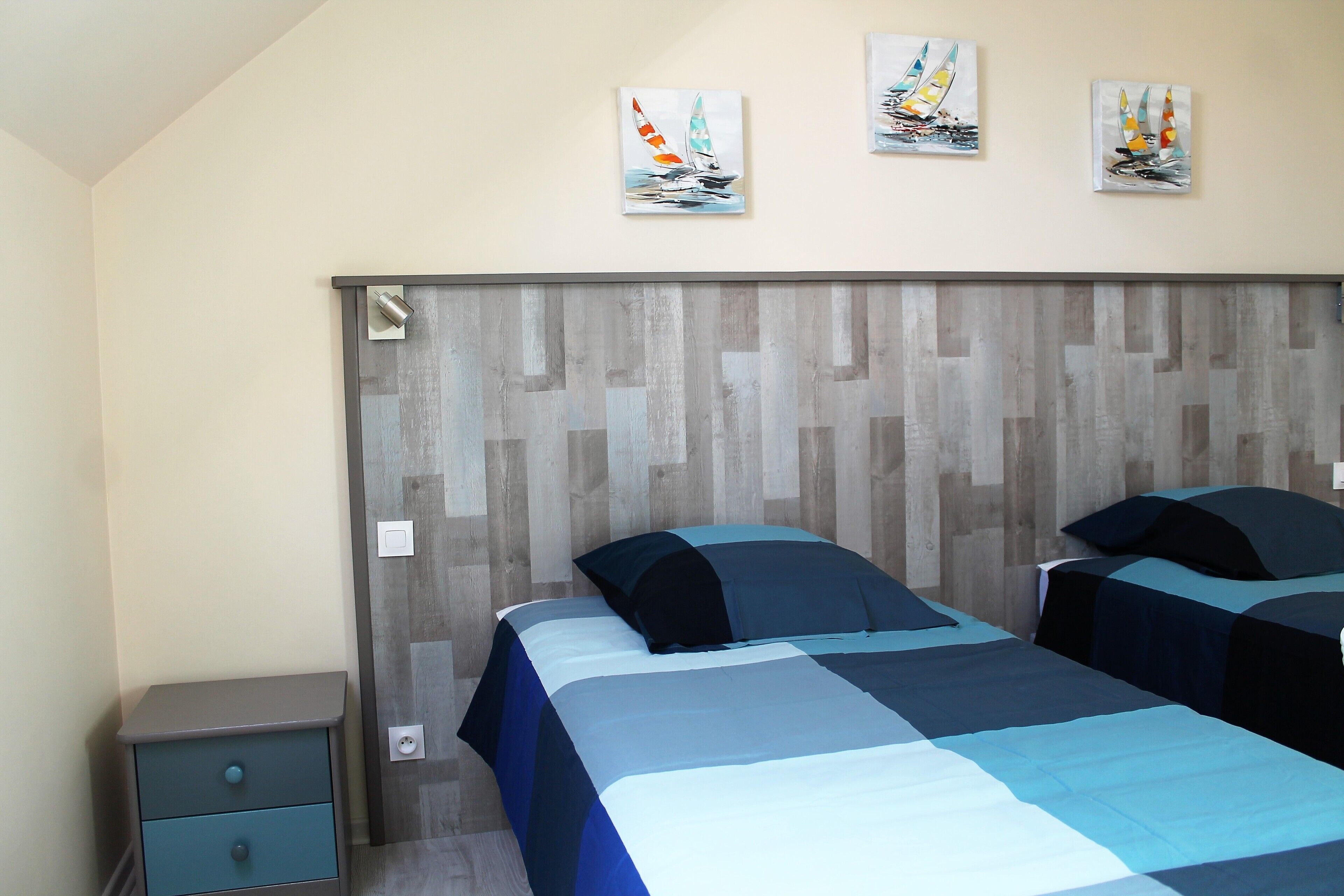 A bedroom with a wooden headboard, two beds with blue bedding, a nightstand, and three sailboat paintings on the wall.