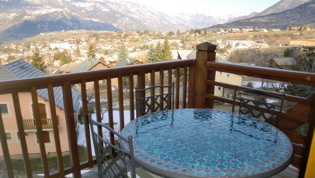 Een balkon met een ronde tafel en stoelen, met uitzicht op een bergachtig landschap met een dorpje beneden.