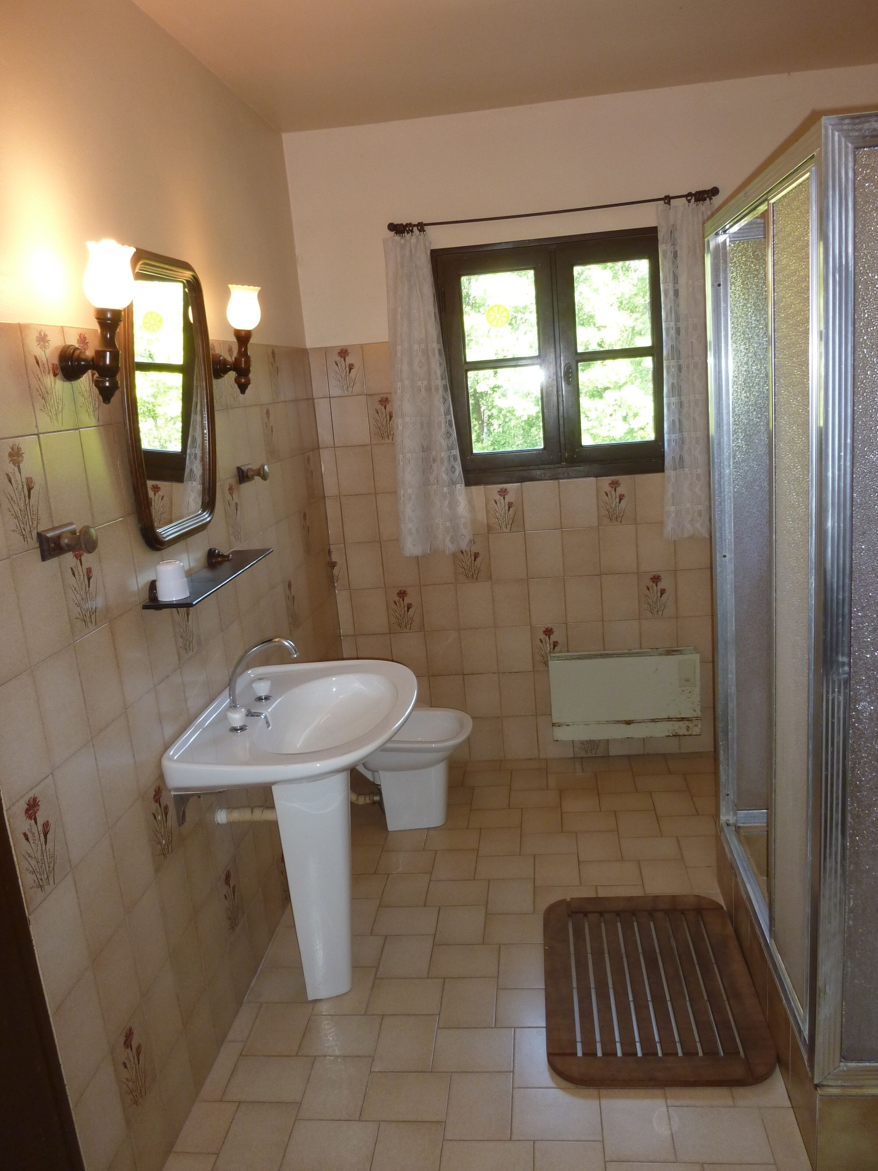Un baño con lavamanos, inodoro y ducha. Hay una ventana con cortinas y las paredes están cubiertas de azulejos con motivos florales.