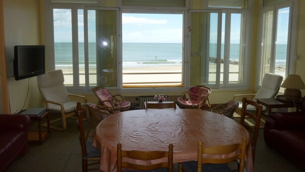 Una habitación con una mesa grande, varias sillas, un televisor y amplios ventanales con vista al mar.