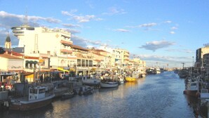 Una marina con barcos amarrados a lo largo de un frente marítimo, edificios con comercios y un cielo despejado.