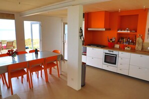 A modern kitchen with white cabinets and orange accents, a dining area with orange chairs and a table, and a view of the outdoors through a large window.