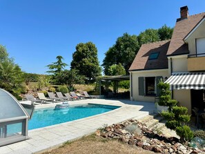 A swimming pool with lounge chairs, a covered patio, and a house with a tiled roof.