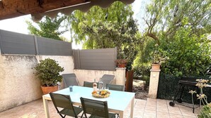 Une terrasse extérieure avec une table blanche et quatre chaises vertes, entourée de plantes en pot et dotée d’un sol carrelé.