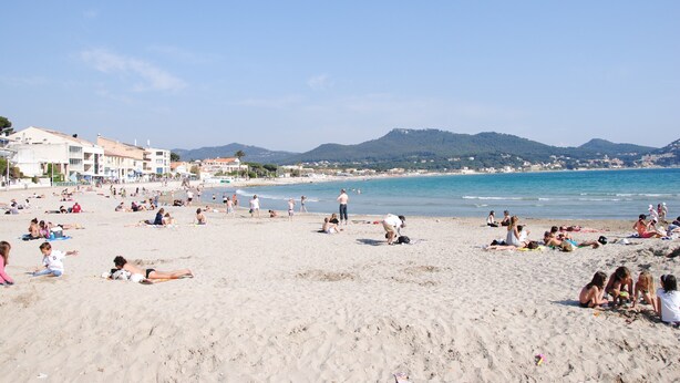 Une plage où des gens se prélassent au soleil et se baignent. On aperçoit des bâtiments au loin et une chaîne de montagnes.