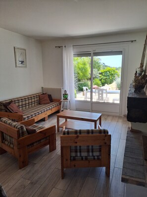 Un soggiorno con divano, due panche in legno, un tavolo e vista su una zona esterna con piante e un tavolo.