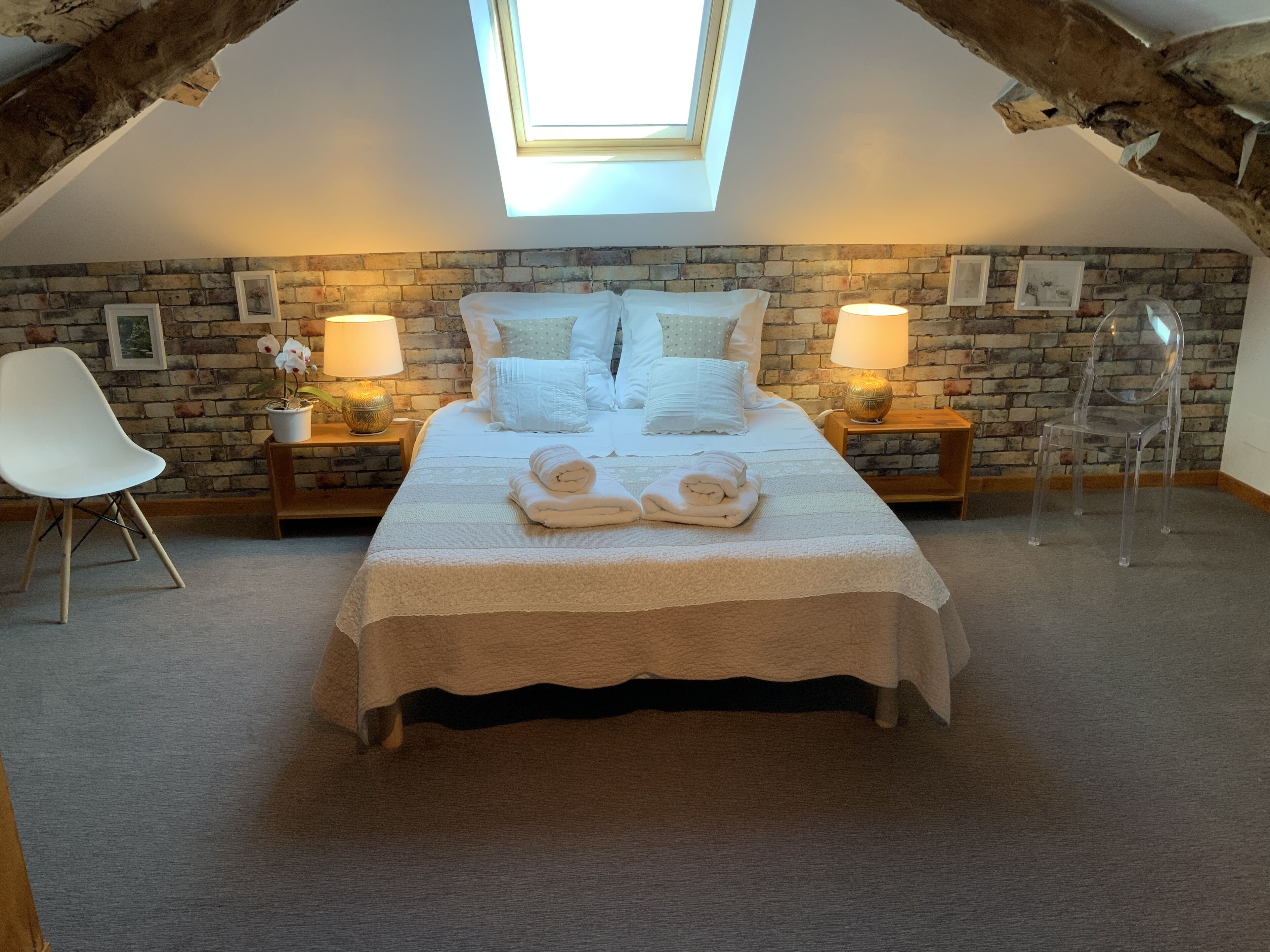 Un dormitorio con techo inclinado, tragaluces, pared de ladrillo, cama con ropa de cama blanca, dos mesitas de noche con lámparas, una silla blanca y una silla de plástico transparente.