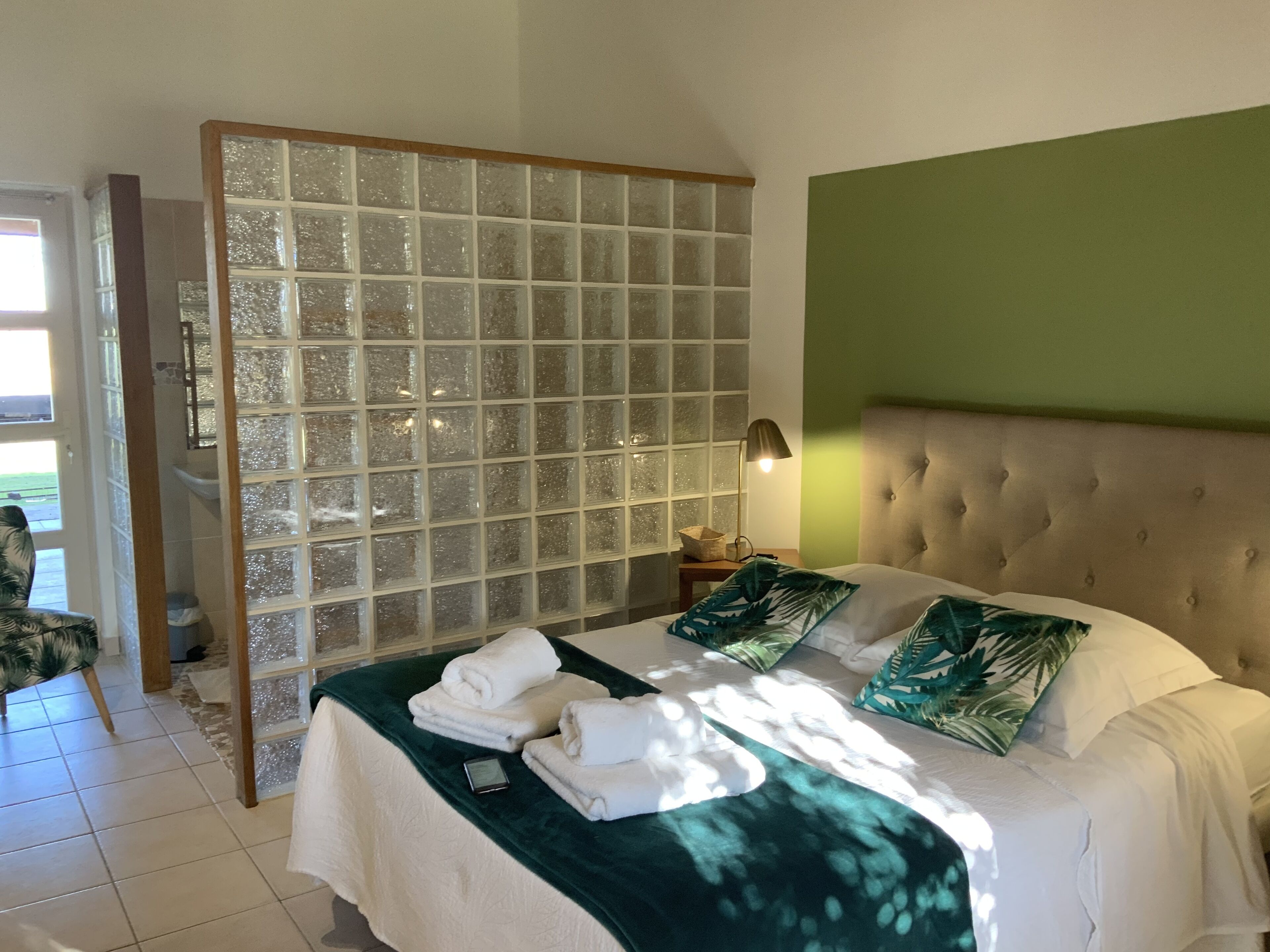 A bedroom with a large bed, a green wall, a glass partition, and a view of the outdoors.
