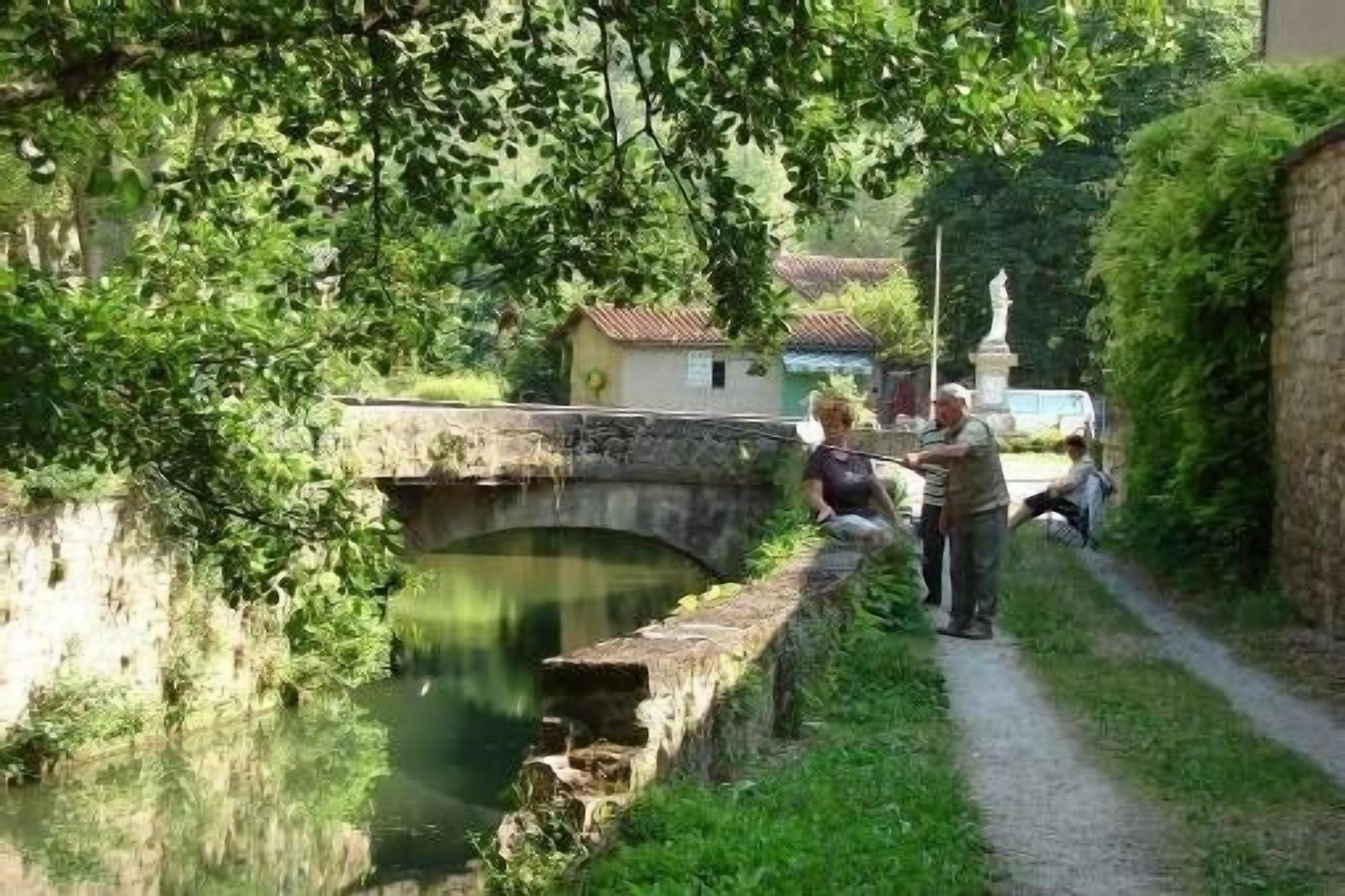A narrow waterway with a stone bridge, lush greenery, and a statue.