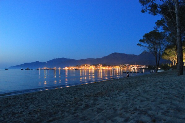 Une plage au crépuscule, avec des eaux calmes reflétant les lumières de la ville, une rive sablonneuse et des montagnes lointaines.