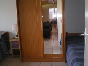 Una habitación con un armario de madera, una cama, una mesita de noche con lámpara y vistas a otra sala con una mesa y sillas de comedor.