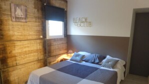 Un dormitorio con pared de madera, una cama con colcha gris y blanca, una mesita de noche con lámpara y un letrero que dice "BEACH CLUB".