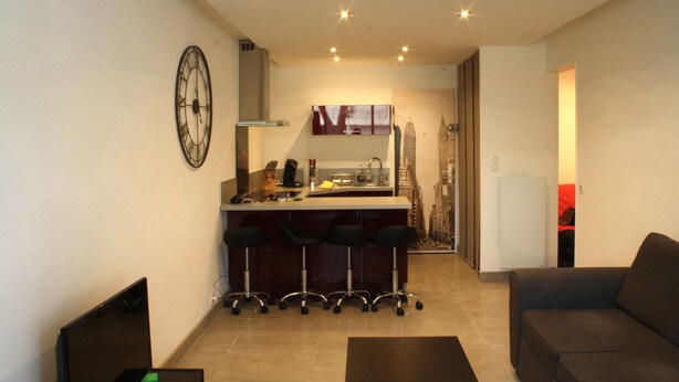 Un espacio habitable compacto con kitchenette, una zona de comedor con taburetes de bar, un sofá y un reloj de pared grande.