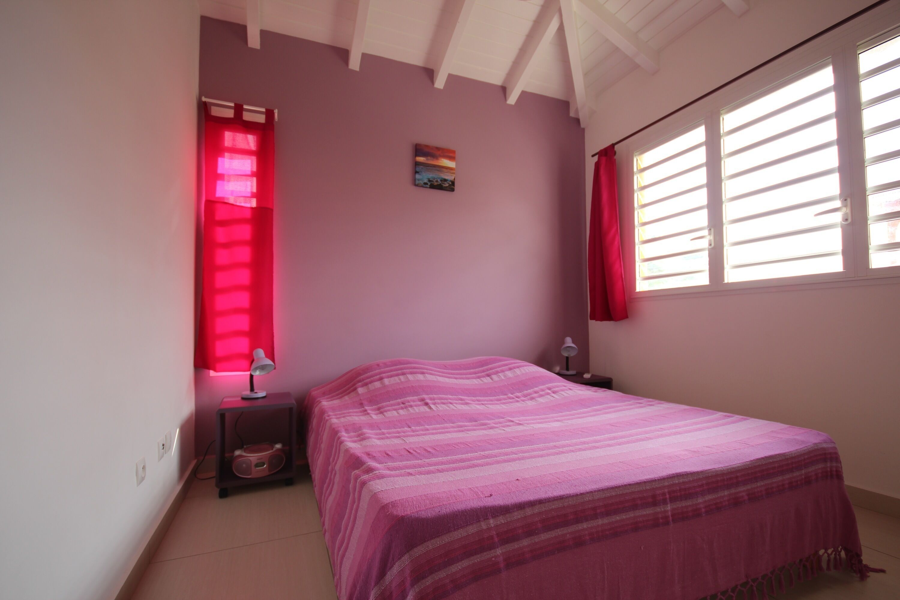 Une chambre à coucher avec un lit, une table de chevet, une lampe, une fenêtre avec des stores et un tableau au mur.