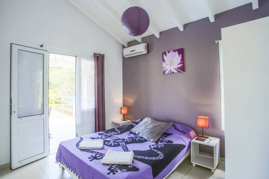A bedroom with a bed, two bedside tables with lamps, a wall-mounted air conditioner, a door, and a window with curtains.