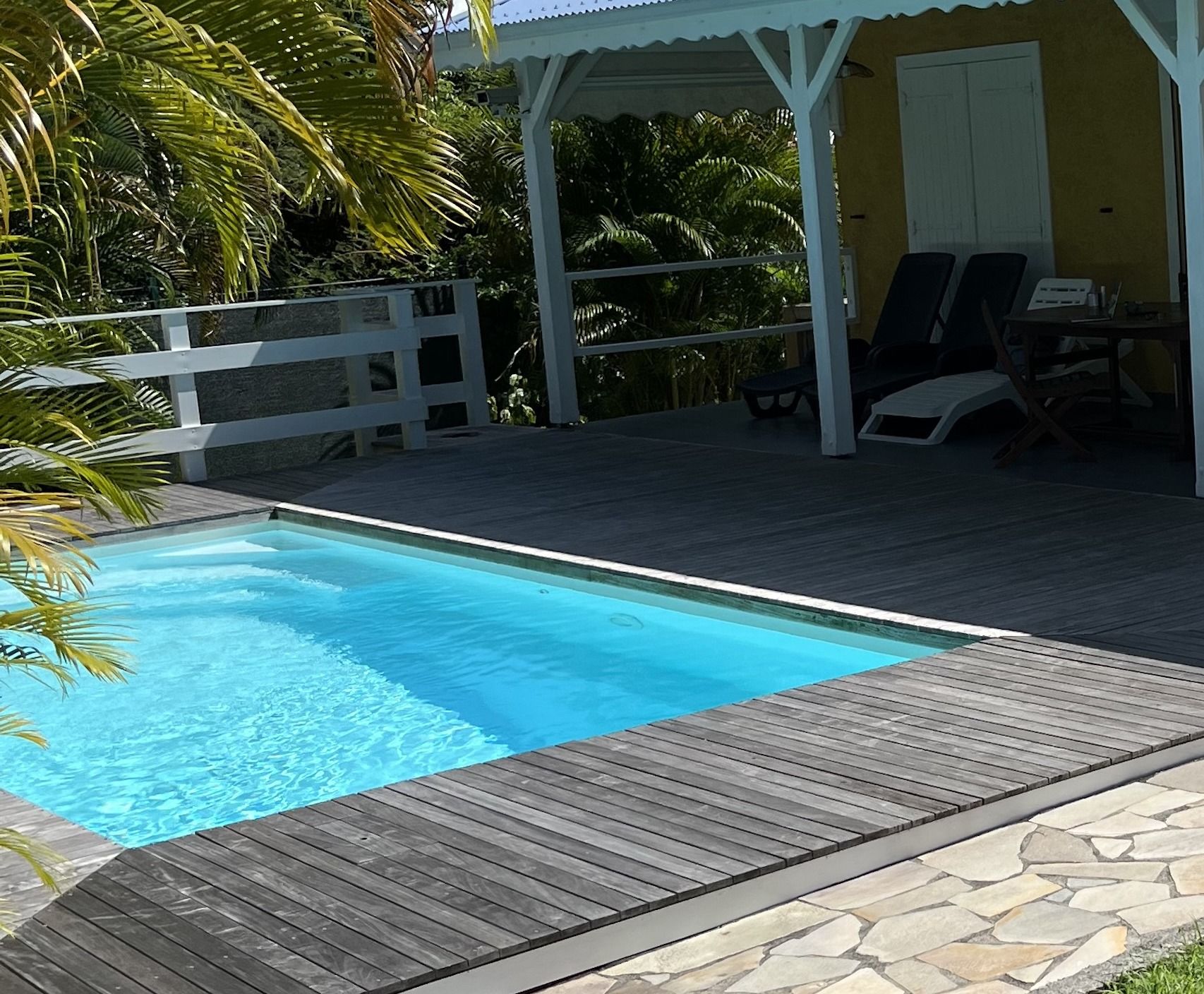 A rectangular swimming pool with a wooden deck surrounding it, a white gazebo, and a small table with chairs.