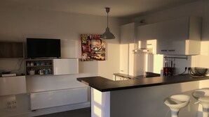 Et moderne køkken med en central ø, hvide skabe, en hængende lampe og et farverigt kunstværk på væggen.