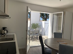 Eine moderne Küche mit Einbauofen und Kaffeemaschine, ein Essbereich mit rundem Tisch und Stühlen, und eine offene Tür zu einer Terrasse im Freien mit Tisch und Stühlen.