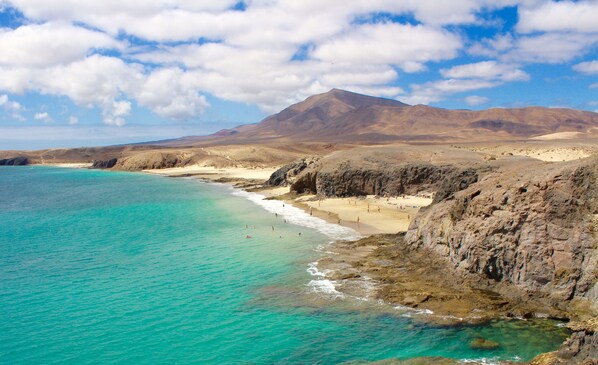 Paisaje costero con aguas turquesas cristalinas, playas arenosas, acantilados rocosos y una montaña al fondo.