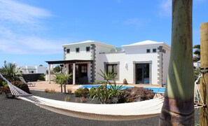 Una casa moderna a due piani con piscina, circondata da piante tropicali e un'amaca.